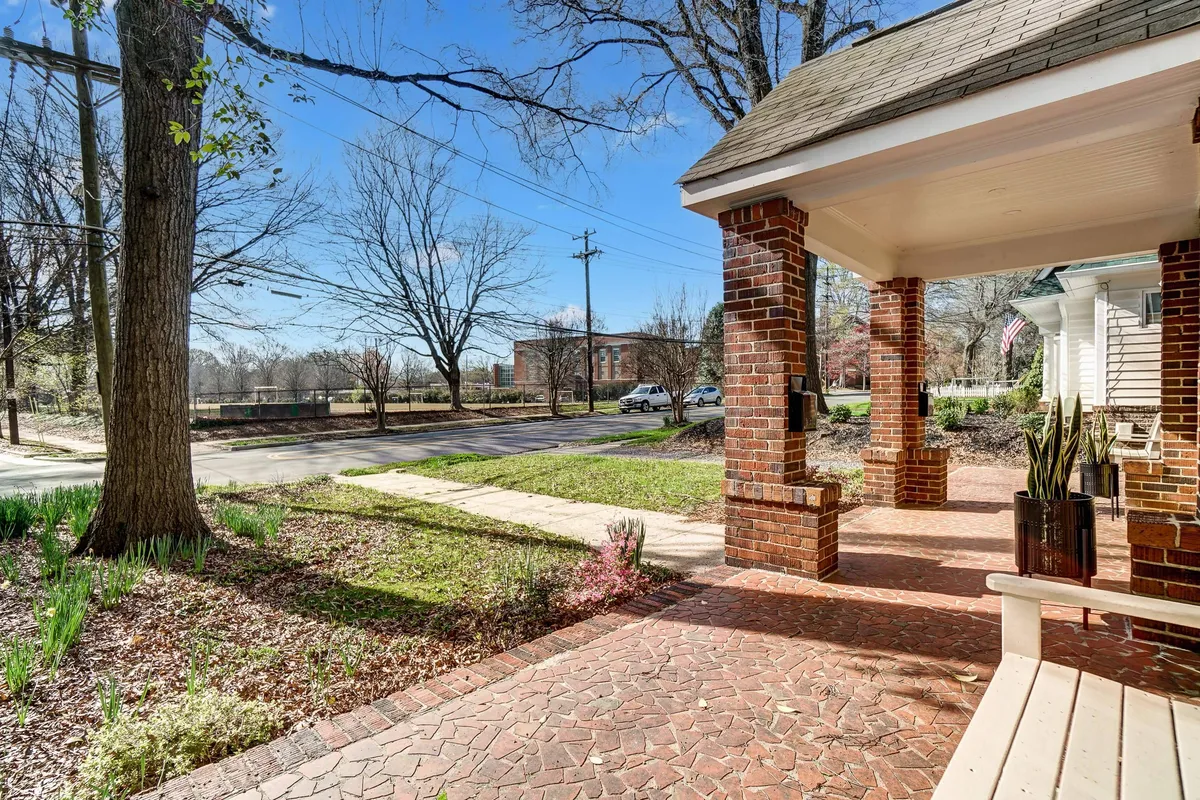 The inviting front porch of a classic red brick duplex, with comfortable seating and seasonal landscaping. The garden pathway leads to the entrance, setting a welcoming tone and highlighting the charm of staying in a historic neighborhood close to the city action.