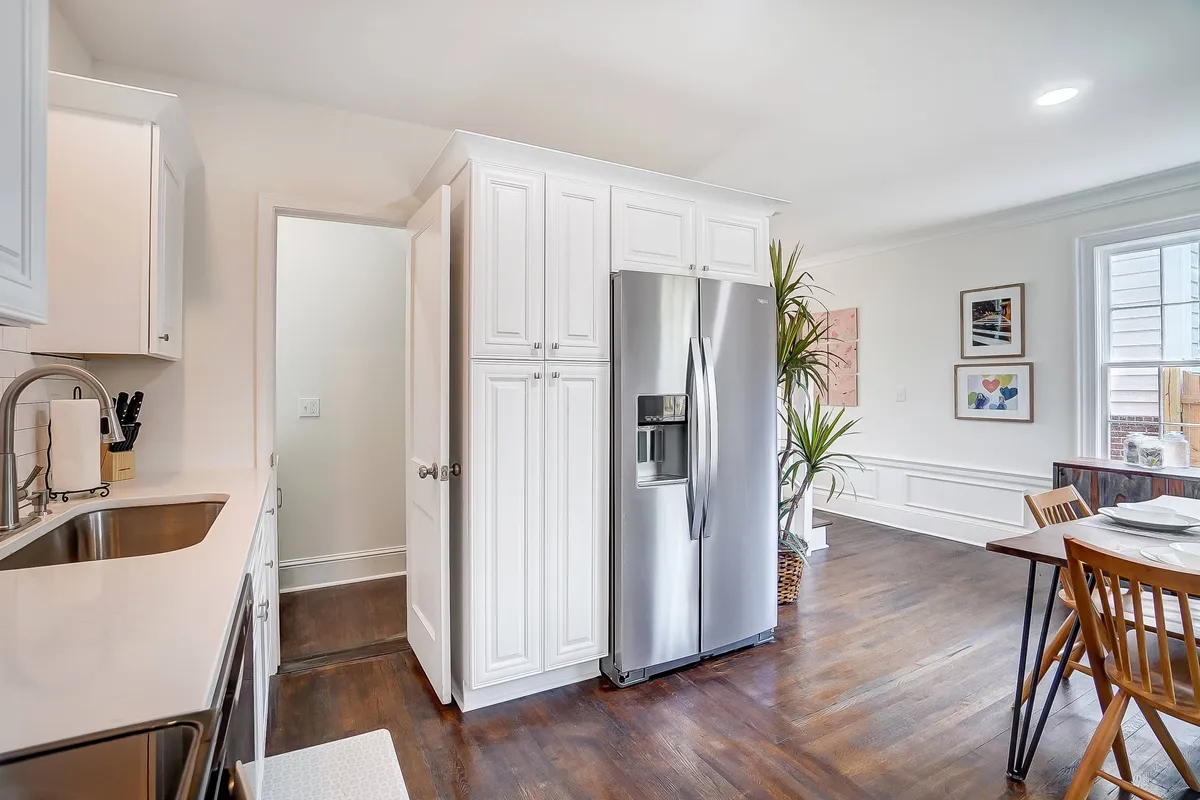 A welcoming view of the kitchen and dining area, featuring a stainless steel refrigerator, kitchen sink, wooden dining table, lush green plants, and framed artwork. This angle highlights the seamless flow between cooking and dining spaces, ideal for casual meals or sharing coffee with friends and family.