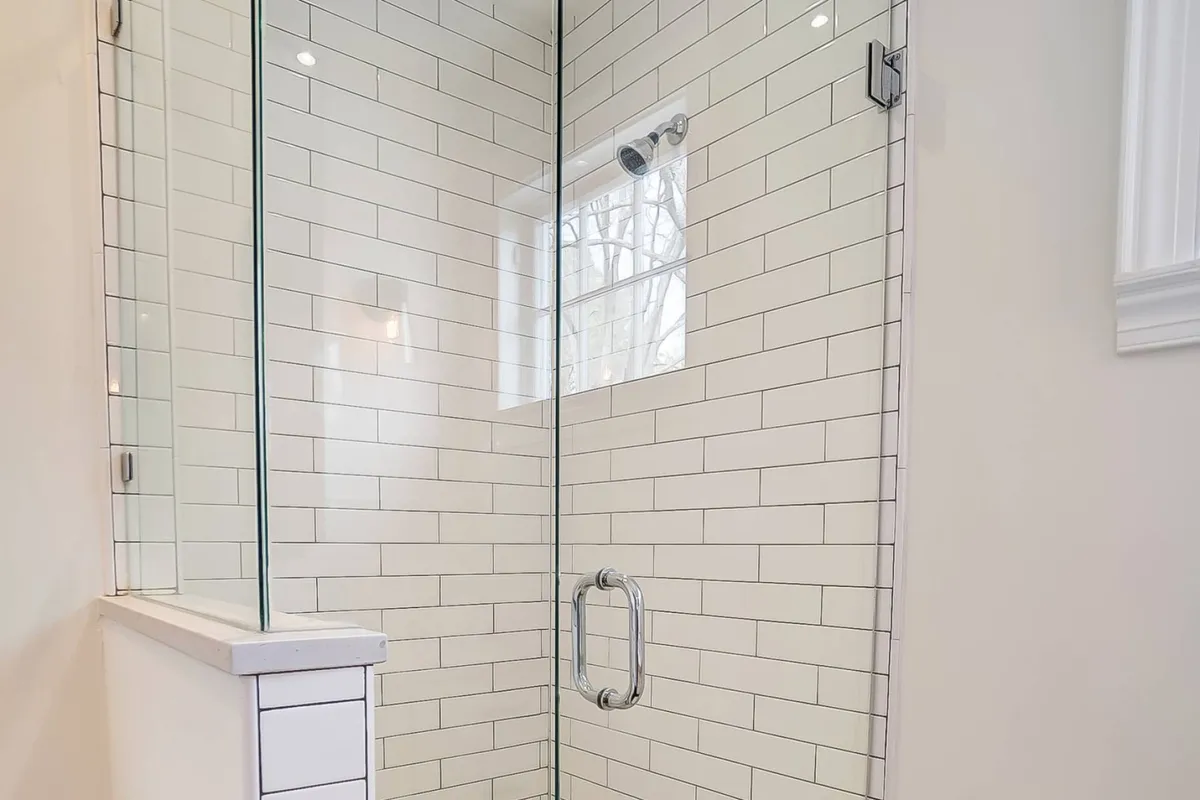 A modern bathroom with a glass shower enclosure, crisp subway tile walls, a sleek showerhead, and a window allowing natural light to filter in. The space is clean, fresh, and styled for relaxation, with towels neatly hung nearby.