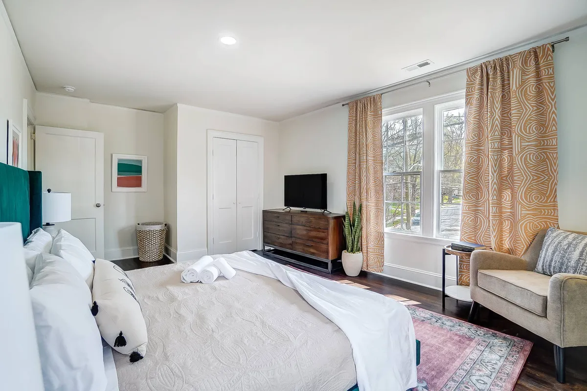 A king bedroom featuring a comfortable bed with decorative pillows, a large window with curtains that lets in generous natural light, a wooden dresser beneath a wall-mounted TV, and an armchair in the corner. The palette is soft and calming, and the room feels spacious and restful.