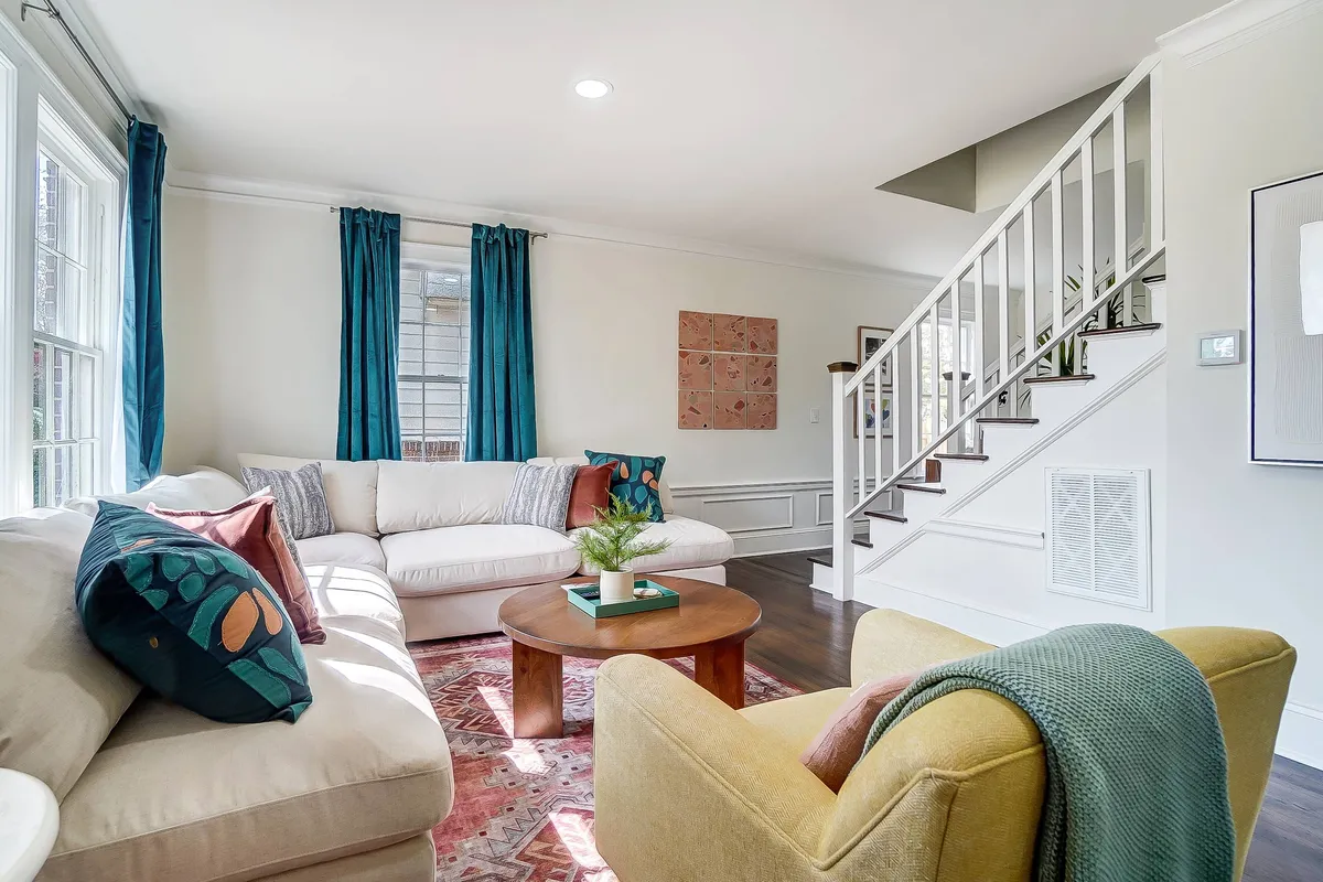 A spacious, welcoming living space showcasing a white sectional sofa, colorful accent chairs, a round coffee table perfect for games or snacks, striking wall art, and a modern staircase that adds visual interest. The room is awash in natural light, enhancing its inviting, familial feel.