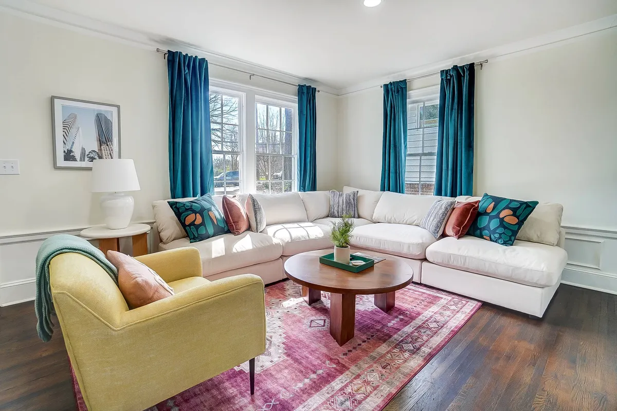 A vibrant living area with a comfortable sofa, colorful decorative pillows, a coffee table set for socializing or playing games, and large, light-filled windows with flowing curtains. The space radiates warmth and features Bohemian-inspired décor, ideal for both relaxation and group gatherings.