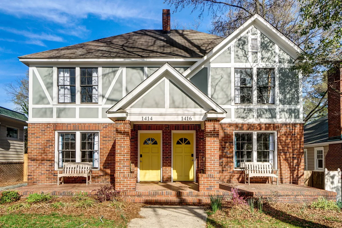 A wide view of the duplex exterior featuring symmetrical yellow front doors, brickwork, white trim, and benches on the porch. The inviting setup suggests social gatherings or a relaxing morning before heading to work.