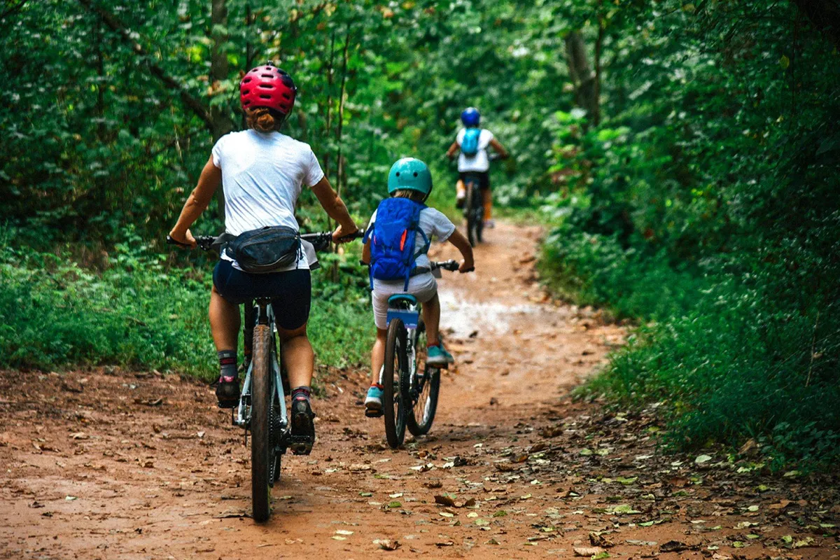 A lively scene of a family biking together along a tree-lined dirt path, with sunlight filtering through the leaves. The natural setting evokes a sense of exploration, adventure, and togetherness.