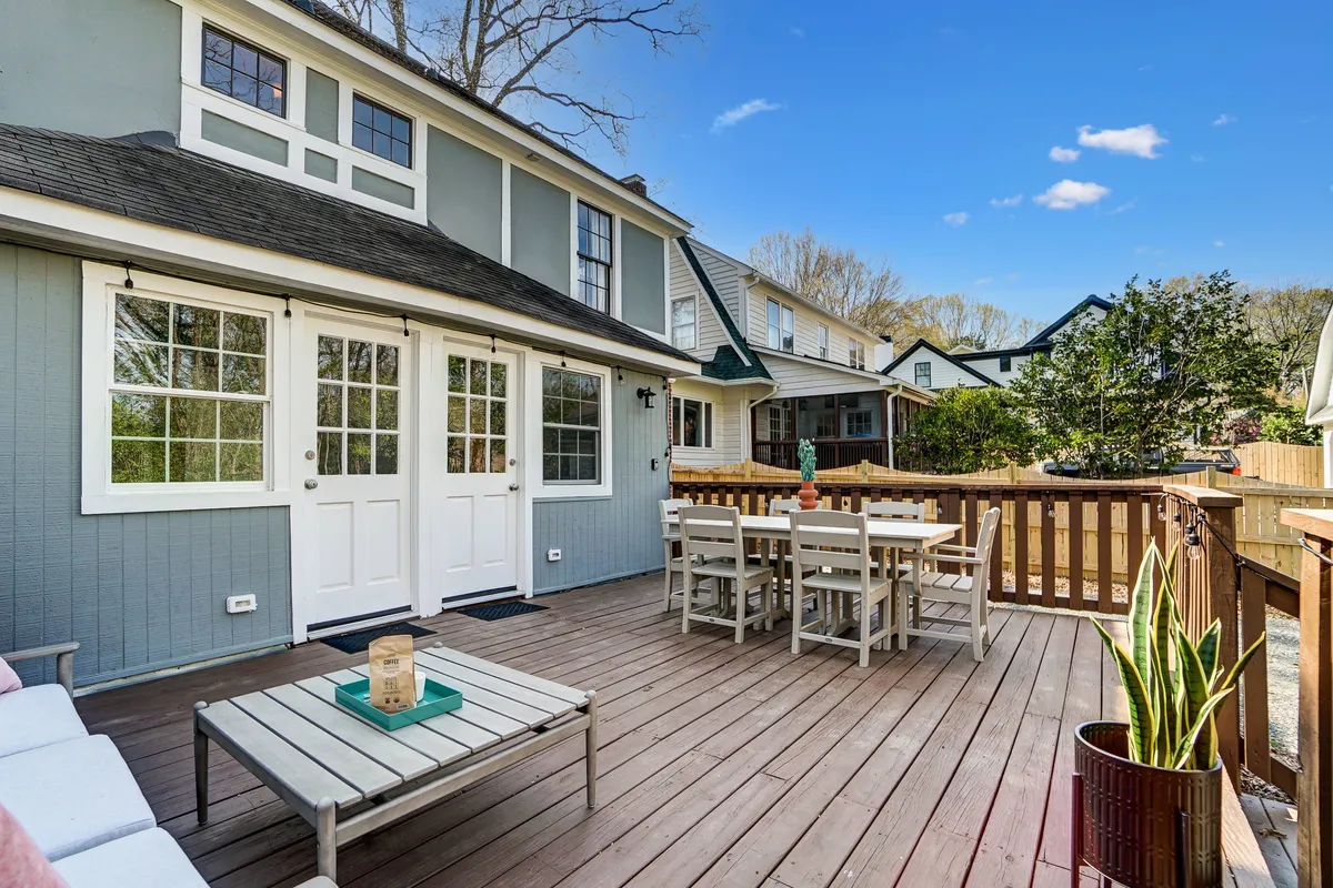 A wooden deck with a dining table and green plant sits just outside double doors, blending seamlessly into the surrounding greenery. The setup is ideal for outdoor dining, social gatherings, and relaxing in a friendly, communal setting.