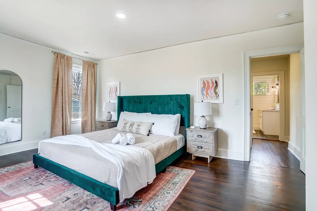 A cozy queen bedroom with a green velvet headboard, crisp linens, colorful area rug, and calming artwork on the walls. Morning light streams in from the window, and the space feels inviting and serene, perfect for unwinding after a long day.
