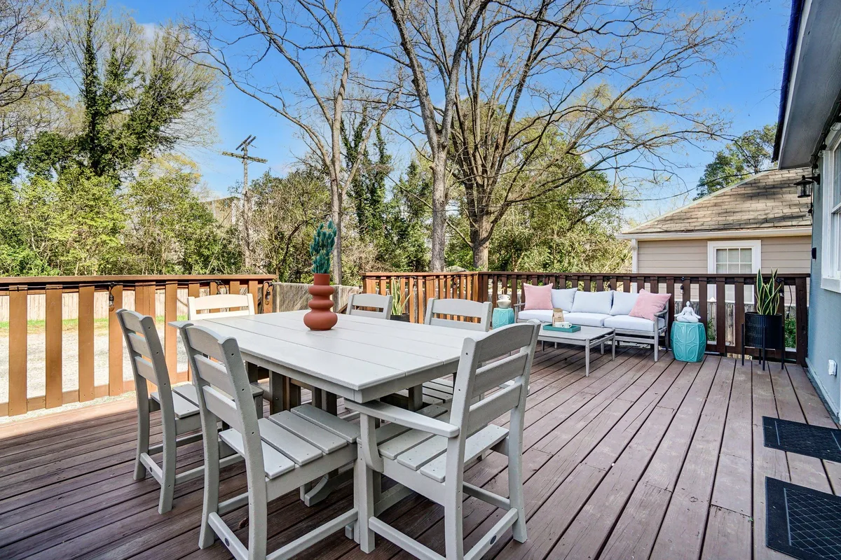 The image showcases a charming outdoor dining area set on a spacious deck, surrounded by lush decorative planters. There's a cozy dining table ideal for gatherings, and inviting outdoor seating perfect for relaxing or enjoying a meal. The atmosphere is casual, warm, and welcoming, suggesting a space ideal for sunset dinners, morning coffee, or celebrating with friends and family.