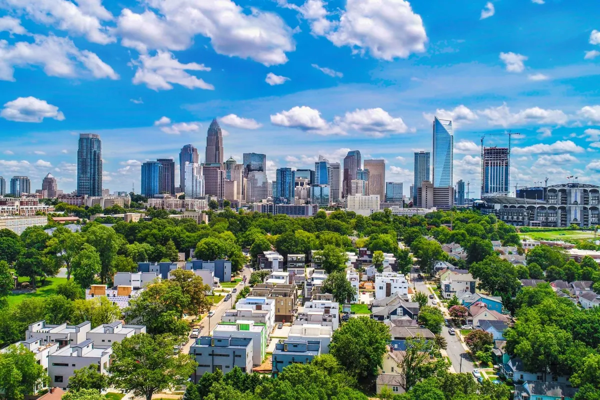 A dynamic view of the Uptown Charlotte city skyline framed by lush green trees and residential homes in the foreground. The composition hints at the proximity to city parks and urban attractions, emphasizing the walkable nature of the neighborhood.